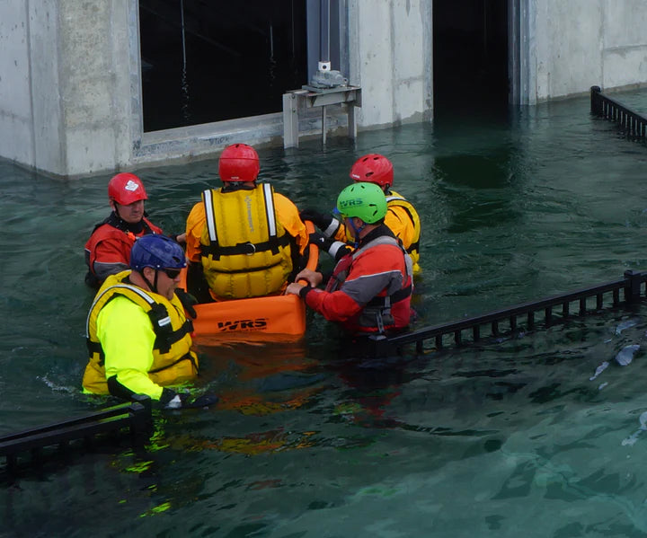 Group of people in safety vests and helmets assisting a person in an orange life raft in water.