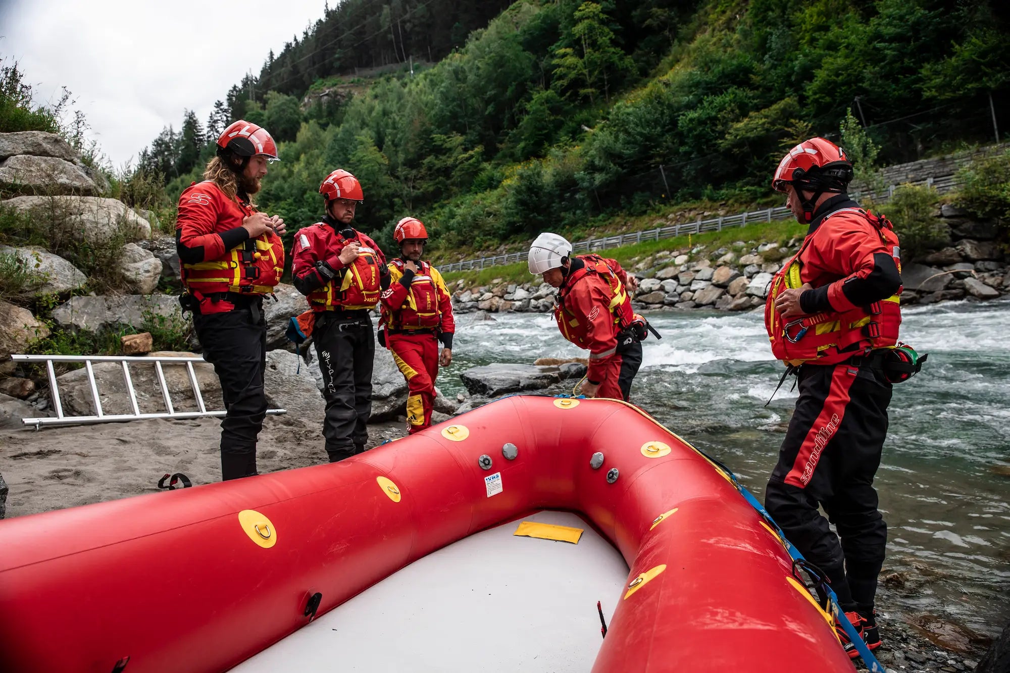 Rescue team in red uniforms and helmets preparing to use a red inflatable raft by a river.