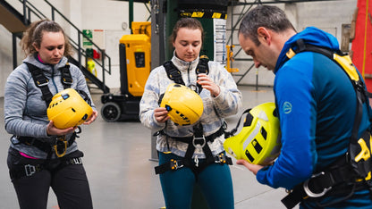 Three people in safety harnesses holding yellow helmets in a warehouse setting.