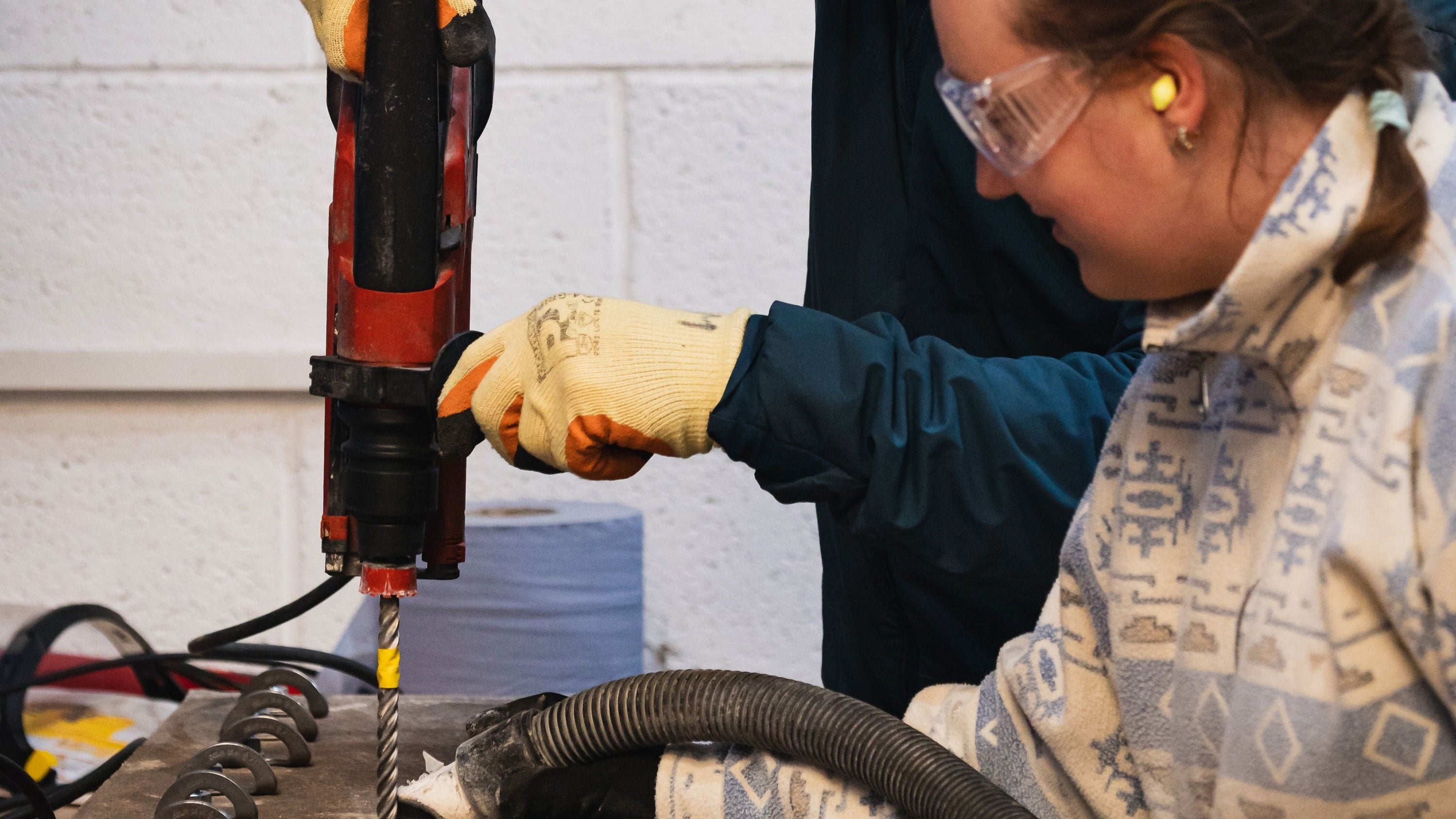 Person using a drill with protective gear against a white wall.