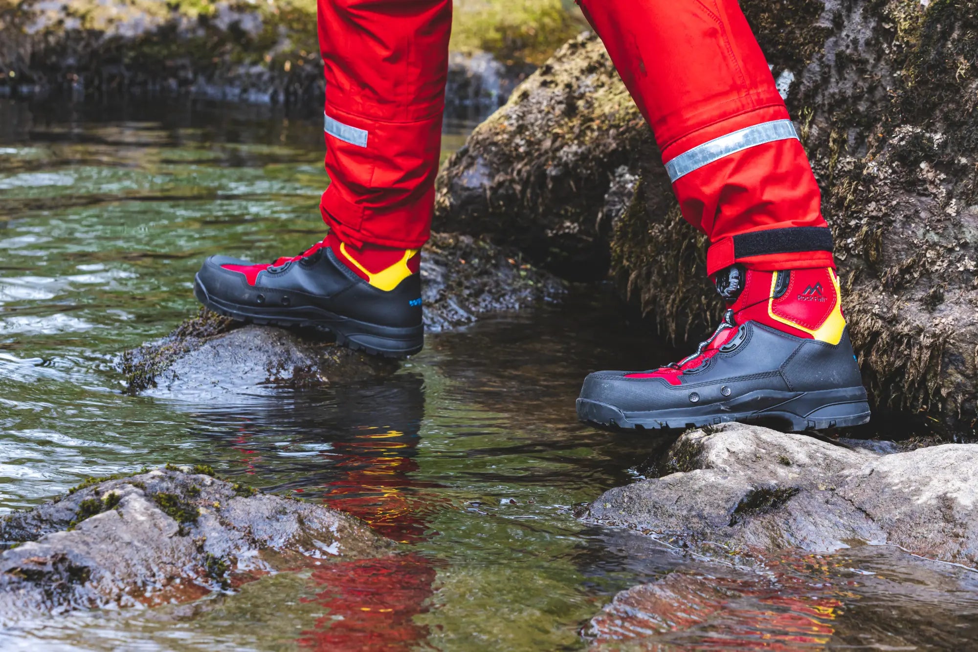 Person wearing red pants and black boots standing on rocks in a stream.