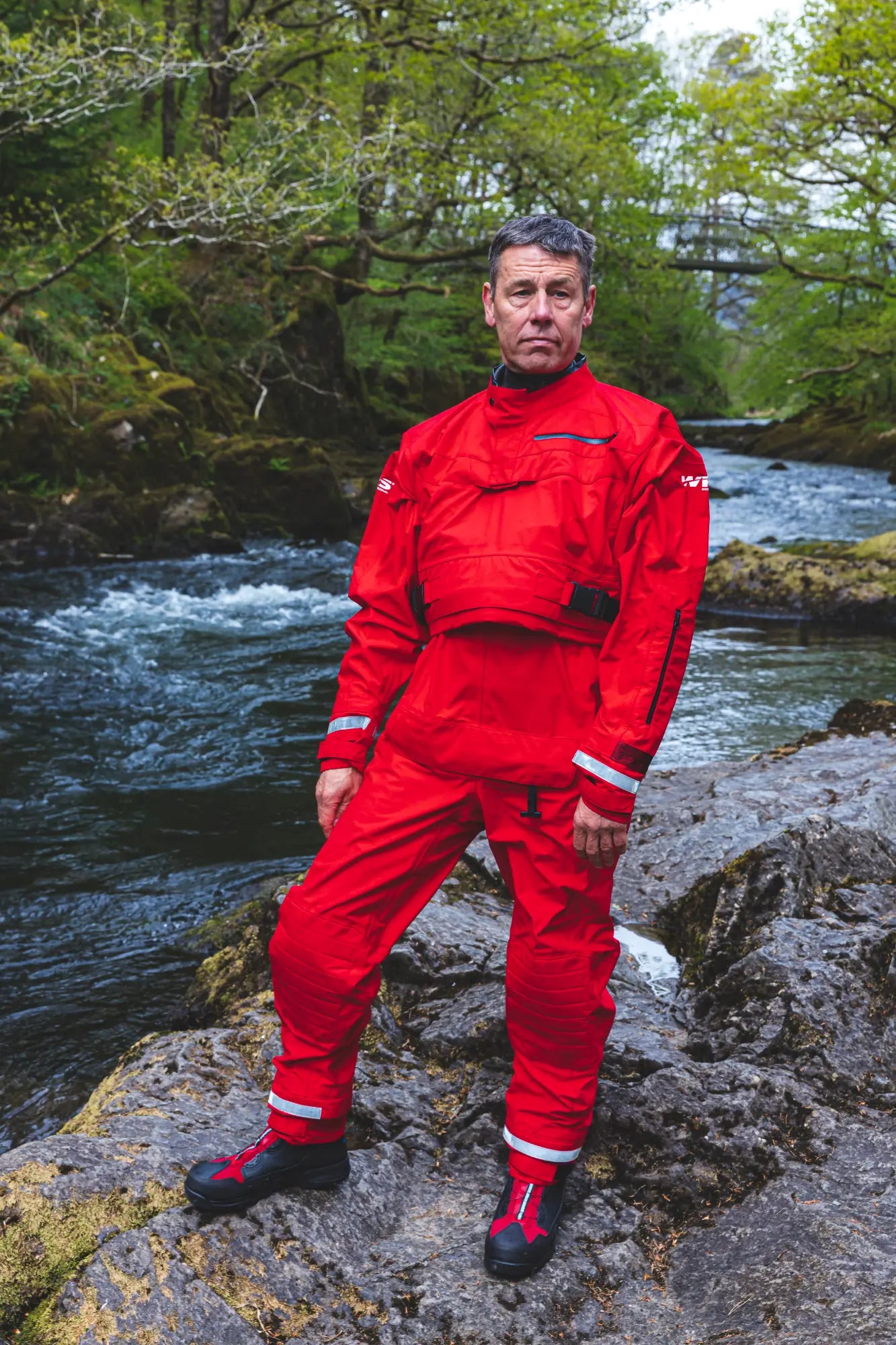 Person wearing a red raincoat and pants standing on rocks by a river with greenery.